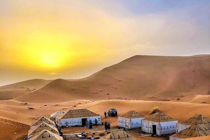 A camel caravan walking across golden dunes under a setting sun in the desert of Morocco.